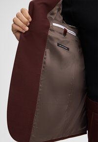 Interno di una giacca marrone bordeaux con fodera liscia marrone, dotata di una tasca laterale con chiusura bianca e etichetta del marchio. Tessuto esterno testurizzato visibile.