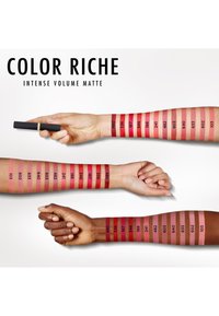 Matowe pomadki Color Riche wyeksponowane na ramionach. Różnorodne odcienie czerwieni i różu, z numerami wskazującymi nazwy kolorów. Tubka pomadki trzymana w powietrzu.