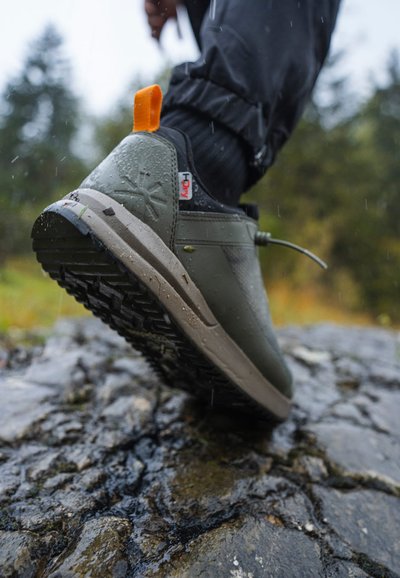 Green waterproof shoe with a textured surface, rubber sole, orange pull tab, and lacing system. Positioned on a wet stone surface.