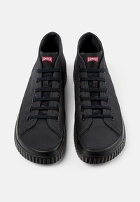 Black canvas sneakers with round toe, textured upper, six eyelets, and rubber sole featuring vertical ridges. Red label on the tongue.