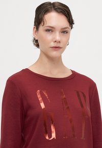 Bordowy sweatshirt z okrągłym dekoltem, z dużym, metalicznym logo "GAP" w kolorze różowego złota z przodu. Miękka tkanina o gładkiej fakturze.