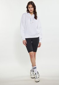 Felpa bianca con coulisse, pantaloncini da ciclismo neri e sneakers bianche con dettagli neri e rossi, abbinate a calzini bianchi a righe.