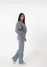 Grijze, gebreide coltrui gecombineerd met hoge, lichtblauwe flared jeans; model met donker haar, draagt doorzichtige hakken.