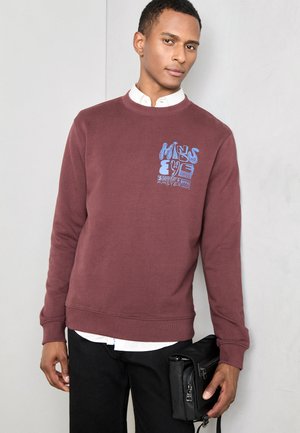 Sudadera color burdeos con texto gráfico azul en el pecho, cuello redondo, puños y dobladillo acanalados. Usada sobre una camisa blanca con cuello y combinada con pantalones negros.