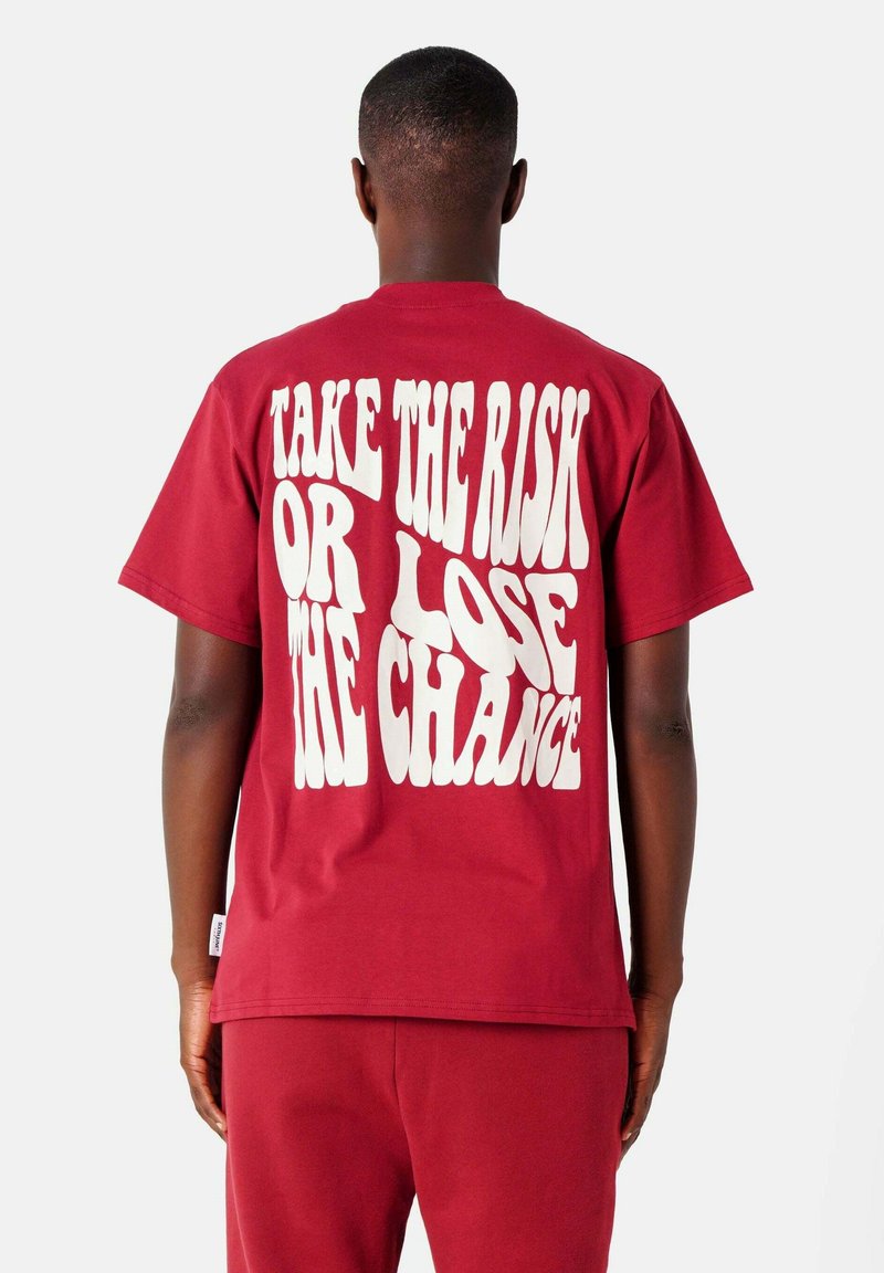 Rode katoenen t-shirt met gedurfde witte tekstontwerp op de achterkant dat zegt "NEEM DE RISICO OF VERLOOP DE KANS." Korte mouwen, ronde hals.