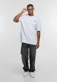 T-shirt grigia oversize con testo stampato sul petto sinistro, abbinata a pantaloni larghi grigi scuri e sneakers bianche. Occhiali con lenti trasparenti.