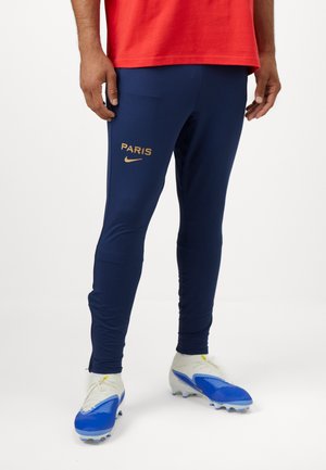 Temno modri atletski hlače s prilagojeno zasnovo, z zlatim napisom "PARIZ" in logotipom Nike na levem stegnu, v kombinaciji z modrimi in belimi nogometnimi čeveljci.