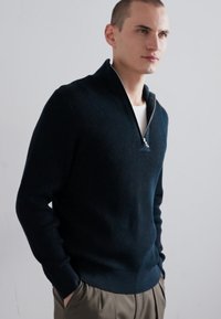 Navy-blauer, gerippter Strickpullover mit einem halben Reißverschluss am Kragen. Besitzt eine strukturierte Oberfläche und eine enganliegende Passform. Wird über einem weißen T-Shirt getragen.