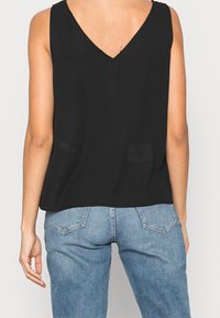 Top senza maniche nero con scollatura a V, texture liscia, vestibilità ampia e dettaglio del logo discreto sul retro. Abbinato a jeans azzurri chiari.