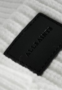 Svart läderetikett med präglad "ALLSAINTS" på strukturerat vitt stickat tyg, som har ett ribbat mönster och en mjuk, fluffig textur.