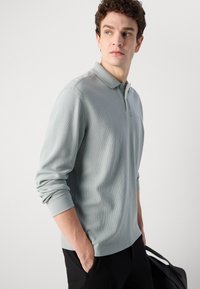 Világosszürke, hosszúujjú pólóing, texturált felülettel, gallérral és megkötött ujjal. Fekete nadrággal és fekete táskával viselve.