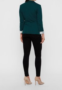 Teal blazer met gestructureerde schouders, slim fit en contrasterende lichtgekleurde manchetten, gecombineerd met zwarte slim jeans en beige hakken.