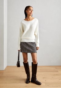 Pull en tricot côtelé blanc, mini-jupe à carreaux noirs et blancs, bottes marron jusqu'au genou, tenant un petit sac noir. Sol en bois, arrière-plan neutre.