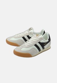 Scarpe sportive bianche e nere realizzate in materiali scamosciati e in tessuto. Presentano una suola in gomma beige, tre strisce nere e un marchio dorato.