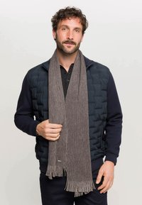 Sciarpa marrone, texturizzata, con dettagli di frange, indossata sopra una giacca blu scuro. Il tessuto liscio e allungato contrasta con il design imbottito.