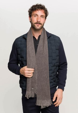 Sciarpa marrone, texturizzata, con dettagli di frange, indossata sopra una giacca blu scuro. Il tessuto liscio e allungato contrasta con il design imbottito.