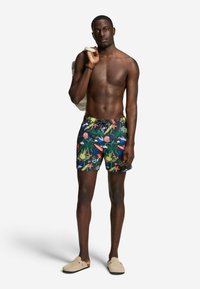 Homme torse nu portant un short de bain tropical coloré, des chaussures beiges faciles à enfiler, portant un sac léger sur une épaule, debout sur un fond uni.