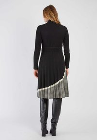 Schwarzes, geripptes Langarmkleid mit einem plissierten Rock, der auf einer Seite ein weißes Hahnentristenmuster aufweist. Kombiniert mit knielangen schwarzen Stiefeln.
