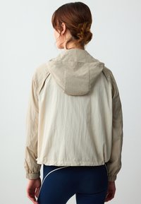 Beige leichte Jacke mit Kapuze, elastischen Bündchen und strukturiertem Finish. Die Rückseite hat einen etwas längeren Saum, der gut zu dunkelblauen Hosen passt.