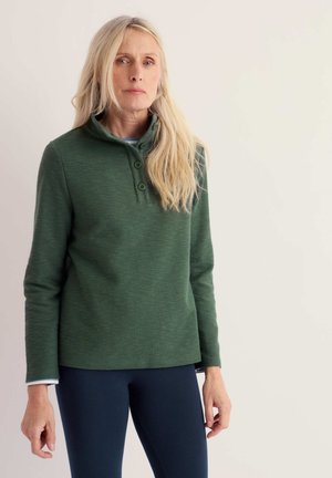Femme aux longs cheveux blonds portant un pull vert à boutons et un pantalon sombre, debout devant un fond clair et uni.