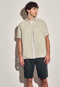 Camisa corta de manga corta en color beige claro con un cierre de botones en la parte delantera y un bolsillo en el pecho, combinada con pantalones cortos de color gris oscuro. Tejido texturizado y casual.