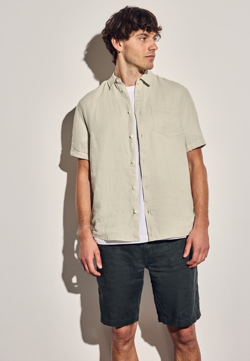 Camisa corta de manga corta en color beige claro con un cierre de botones en la parte delantera y un bolsillo en el pecho, combinada con pantalones cortos de color gris oscuro. Tejido texturizado y casual.