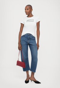 Hvit tettsittende t-skjorte med "ARMANI EXCHANGE" tekst, blå baggy denimjeans, svarte spisse flats, og en liten rød håndveske.