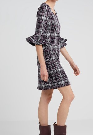 Woman wearing a knee-length purple and white patterned dress with ruffled three-quarter sleeves and brown ankle boots, standing sideways.