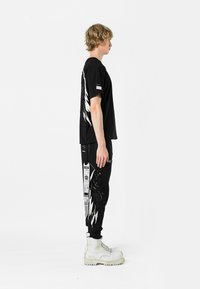 Camiseta gráfica negra con diseño de alas blancas en la parte trasera, combinada con pantalones jogger negros con detalles impresos y acentos blancos. Botas blancas.