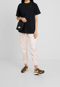 Svart t-shirt, ljusrosa satin-joggers, bruna sneakers med vit accent och svart sula, samt en liten svart crossbody-väska.