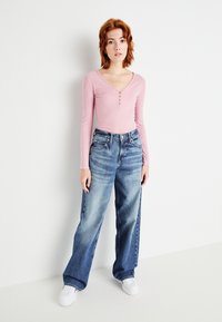 Lys rosa ribbet langermet topp med V-hals og knappedetalj, parret med løse blå denimjeans og hvite joggesko.