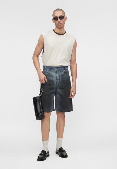 Άντρας που φοράει λευκό αμάνικο τοπ, μαύρα τζιν σορτς με ψηλή μέση και διαβάθμιση, μαύρα μοκασίνια με λευκές κάλτσες, γυαλιά ηλίου και κρατάει μαύρη τσάντα.