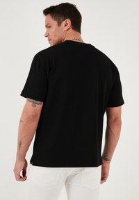 Juodas medvilnės marškinėliai su laisvu kirpimu, trumpomis rankovėmis ir apvaliu iškirpimu. Matomos tatuiruotės ant rankos; modelis vilki baltas kelnes.