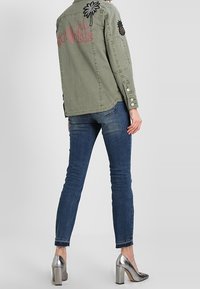 Oliivivihreä farkkutakki, jossa on brodeerattu palmupuu ja ananaksen yksityiskohtia, yhdistettynä istuviin sinisiin farkkuihin ja metallinhohtoisiin hopeakenkiin.