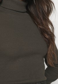 Un haut à col roulé côtelé vert olive foncé, silhouette ajustée, tissu texturé, col haut, manches longues, détails de couture subtils.