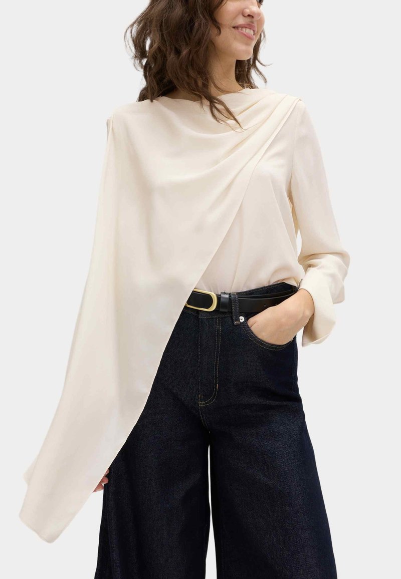 Blusa color crema con diseño asimétrico y caída elegante. Mangas largas, textura suave. Combinada con pantalones de mezclilla oscuros de pierna ancha y cintura alta, acompañados de un cinturón negro.