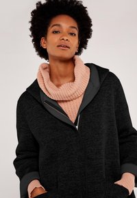 Svart zip-up oversized jacka med en slät textur, utrustad med huva och kontrasterande rosa stickad cowl-hals. Dekorativ dragkedjedetalj.