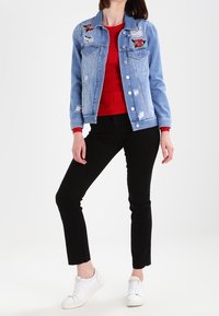 Světle modrá džínová bunda s vyšívanými nášivkami, s opotřebovanými detaily a kovovými knoflíky, spárovaná s červeným žebrovaným svetrem a černými kalhotami.