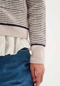 Knit sweater in cream with navy horizontal stripes, ribbed cuffs, layered over a sheer white blouse with textured dots, paired with blue denim.