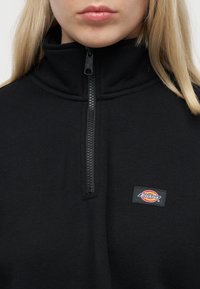 Κλείσιμο εικόνας ενός ατόμου που φοράει μαύρο πουλόβερ με φερμουάρ, ψηλό γιακά και διακριτικό λογότυπο Dickies στο στήθος.