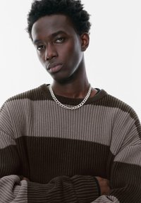 Pull en tricot rayé vert et marron avec une texture côtelée. Collier en chaîne argentée et un sous-vêtement noir visible sous le pull.