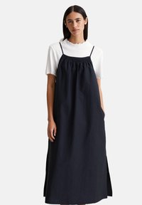 Marineblaues Leinenkleid mit verstellbaren Trägern, gerafftem Brustbereich und Seitentaschen, das über einem weißen, gerippten Oberteil mit gewelltem Saum getragen wird.