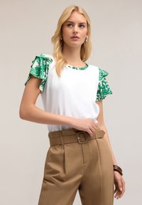 Top bianco a maniche corte con ruches verdi a fantasia e scollatura abbinata, abbinato a pantaloni marroni a vita alta e una cintura.