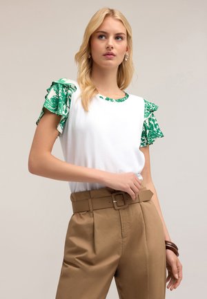 Top bianco a maniche corte con ruches verdi a fantasia e scollatura abbinata, abbinato a pantaloni marroni a vita alta e una cintura.