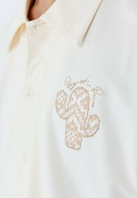 Camisa de cuello en tejido crema claro que presenta un diseño texturizado de cactus bordado en color beige con un patrón a cuadros y detalles de costura.