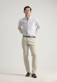 Camisa de botón a rayas blancas y azules, pantalones beige claros, cinturón marrón oscuro y mocasines marrones; pose de pie con las manos detrás de la espalda.