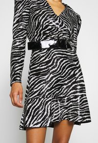 Personne portant une robe zébrée noire et blanche à paillettes, à manches longues, avec une large ceinture noire brillante et une jupe évasée.