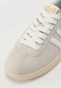 Hellgraue Wildledersneaker mit weißen flachen Schnürsenkeln, sandfarbener Gummisohle und einem kleinen goldenen Emblem auf der Zunge. Verfügt über ein gesticktes Design.