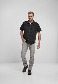 Svart kortermet skjorte med knapper og to brystlommer, grå slitte jeans og svarte solide sko. Nøytral bakgrunn.