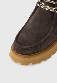 Bruine suede laars met een dikke tan rubberen zool en geweven veters met een houndstooth patroon, die de casual, robuuste ontwerpelementen benadrukken.